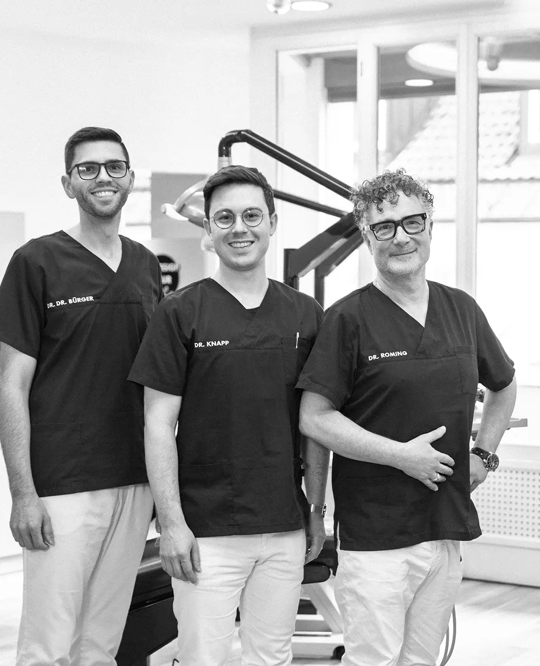 Drei Männer in medizinischer Arbeitskleidung stehen nebeneinander in einem Behandlungsraum und lächeln in die Kamera