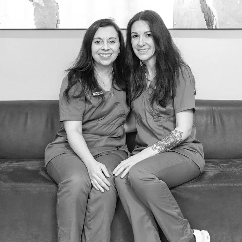 Zwei Frauen in medizinischer Arbeitskleidung sitzen eng nebeneinander auf einem Sofa und lächeln in die Kamera