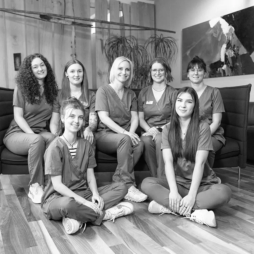 Sieben Frauen in medizinischer Arbeitskleidung sitzen und knien gemeinsam vor einem Sofa und lächeln in einem modern eingerichteten Praxisraum mit Pflanzen und Wandbild