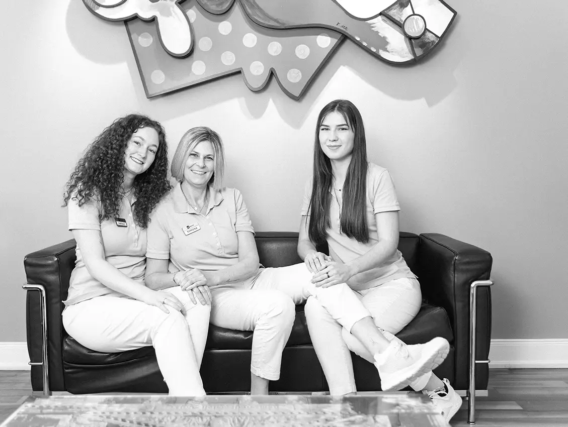 Drei Frauen in medizinischer Arbeitskleidung sitzen gemeinsam auf einem Sofa und lächeln in einem Praxisraum mit dekorativer Wandgestaltung