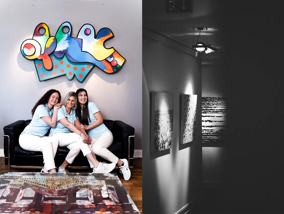 Drei lächelnde Frauen in einheitlicher Kleidung sitzen auf einem Sofa unter einem bunten Kunstwerk in einer Praxis während daneben ein Flur mit beleuchteten Bildern zu sehen ist