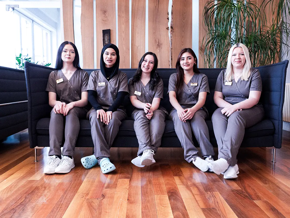 Fünf junge Frauen in medizinischer Arbeitskleidung sitzen nebeneinander auf einem Sofa und schauen freundlich in die Kamera in einem modern eingerichteten Raum mit Holz- und Pflanzenakzenten