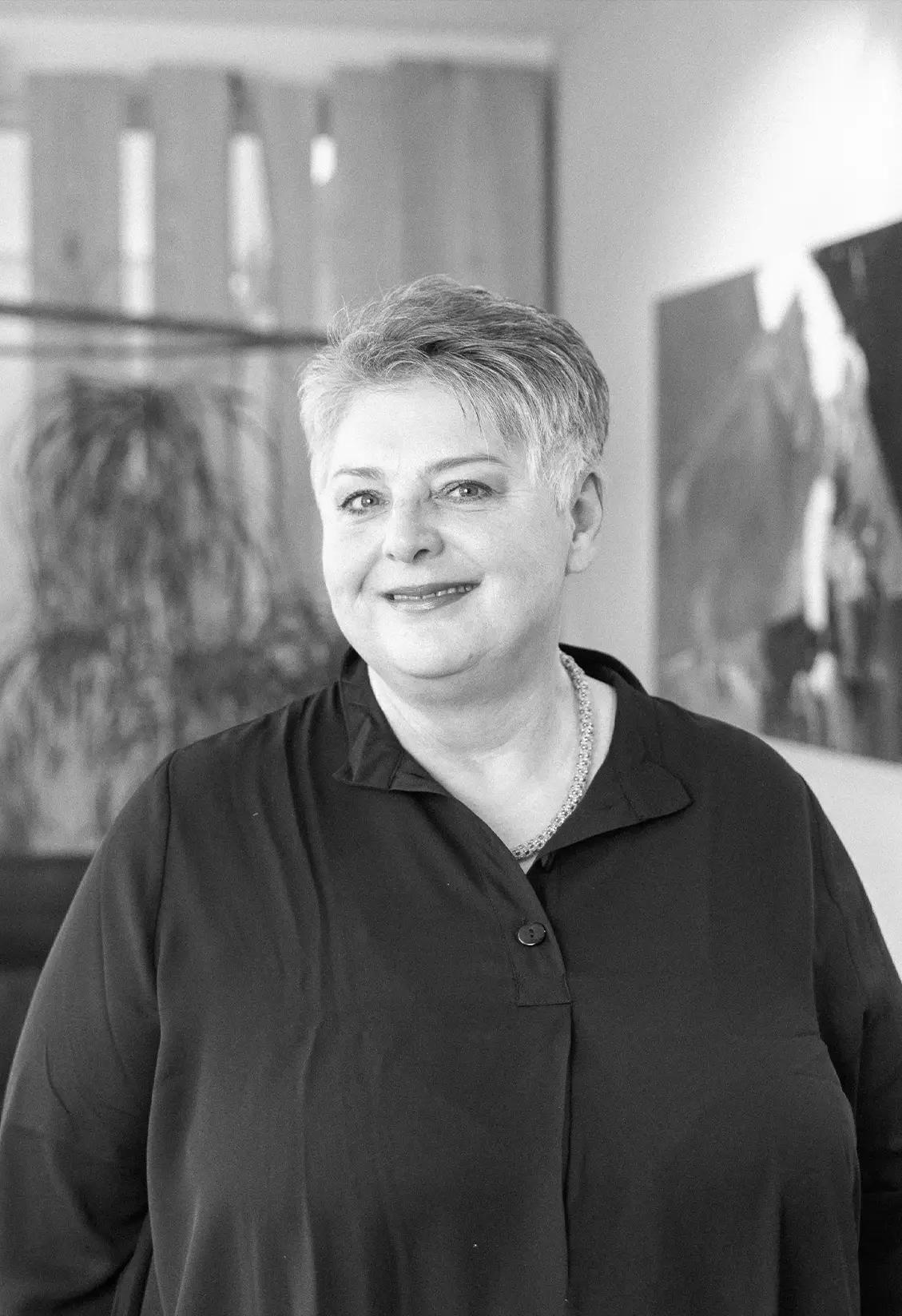 Schwarzweißporträt einer lächelnden Frau mit kurzem Haar in dunkler Bluse vor unscharfem Innenraum mit Pflanze und Wandbild