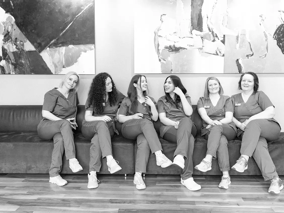 Sechs Frauen in medizinischer Arbeitskleidung sitzen nebeneinander auf einem Sofa, unterhalten sich und lachen gemeinsam in einem Praxisraum mit großen Wandbildern