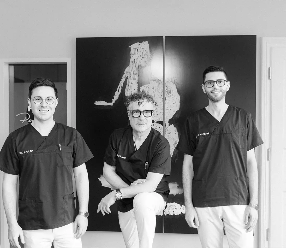 Drei Männer in medizinischer Kleidung stehen vor großformatigen Kunstwerken in einer Praxis und lächeln in die Kamera